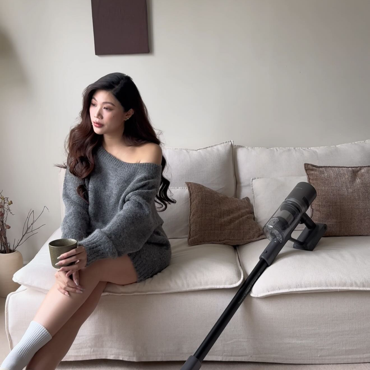 Woman sitting on a couch holding a cup with a vacuum cleaner beside her
